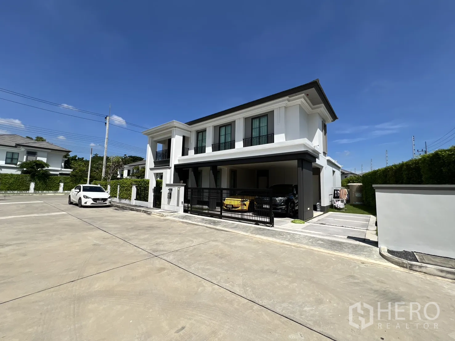 Detached House for rent or sale in Mueang Pathum Thani, Pathum Thani - Angled street view of the white two-storey house with wide driveway and covered parking.