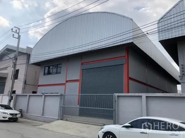 Warehouse for rent in Nonthaburi - Street view of a gated warehouse featuring a wide roller shutter, metal cladding and on-site parking along a quiet access road.