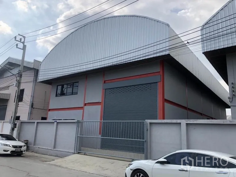 Warehouse for rent in Nonthaburi - Street view of a gated warehouse featuring a wide roller shutter, metal cladding and on-site parking along a quiet access road.