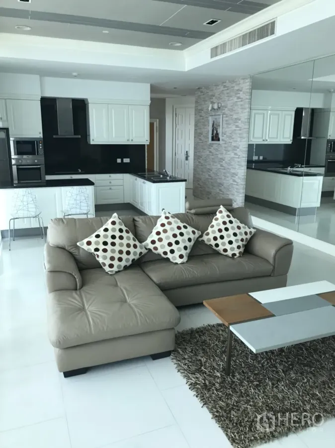 Condominium for rent in Watthana, Bangkok - Open-plan living room with beige leather sofa, shag rug, and white modern kitchen with black backsplash.