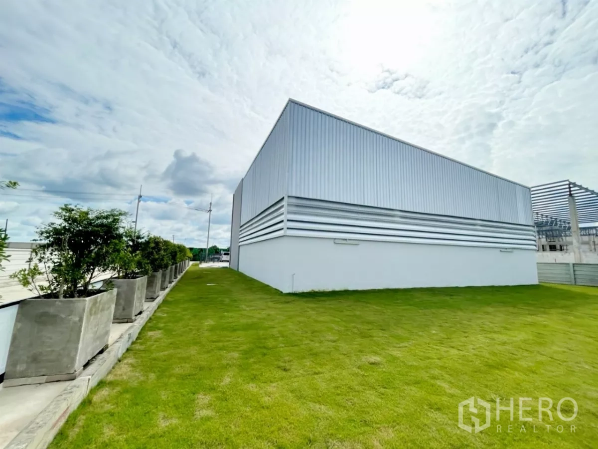 Warehouse for sale in Lam Luk Ka, Pathum Thani - Green lawn and a row of planters along the side of a metal-clad warehouse.