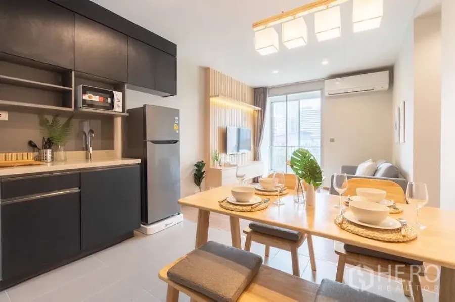 Condominium for rent or sale in Watthana, Bangkok - Open-plan kitchen and dining with black cabinetry, a wooden table set, and a bright living area leading to a balcony.