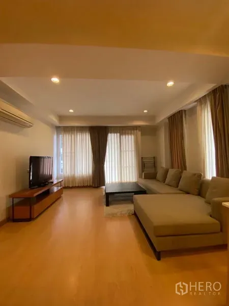 Condominium for rent in Watthana, Bangkok - Bright living room with an L-shaped sofa, TV console, wood floors, and floor-to-ceiling curtains.