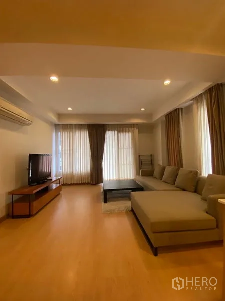 Condominium for rent in Watthana, Bangkok - Bright living room with an L-shaped sofa, TV console, wood floors, and floor-to-ceiling curtains.