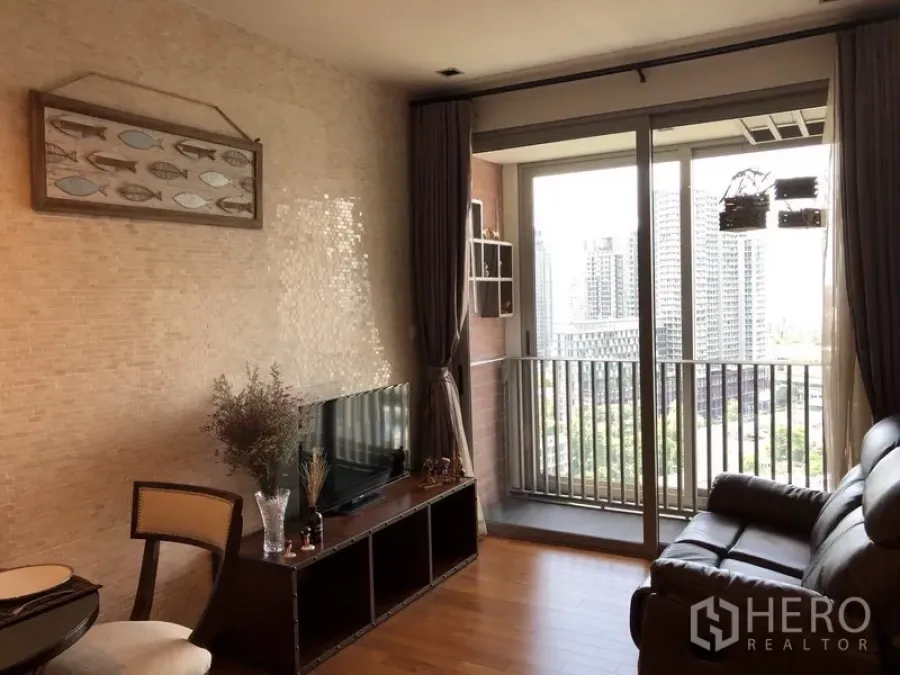 Condominium for rent in Khlong Toei, Bangkok - Bright living room with leather sofa, TV console and sliding doors opening to a balcony with city skyline view.