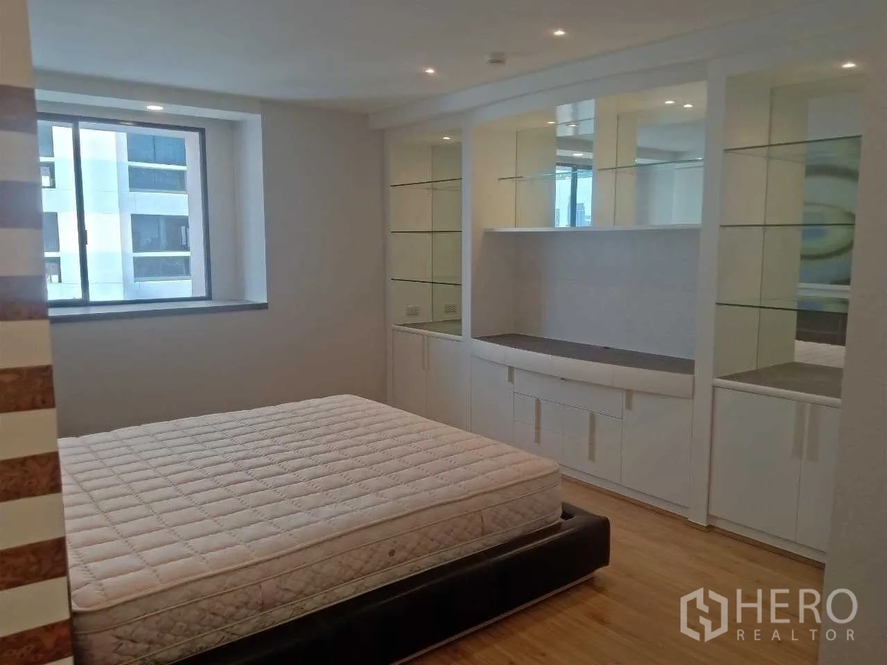 Condominium for rent in Khlong Toei, Bangkok - Bedroom with built-in white wardrobes and glass shelving beside a window.