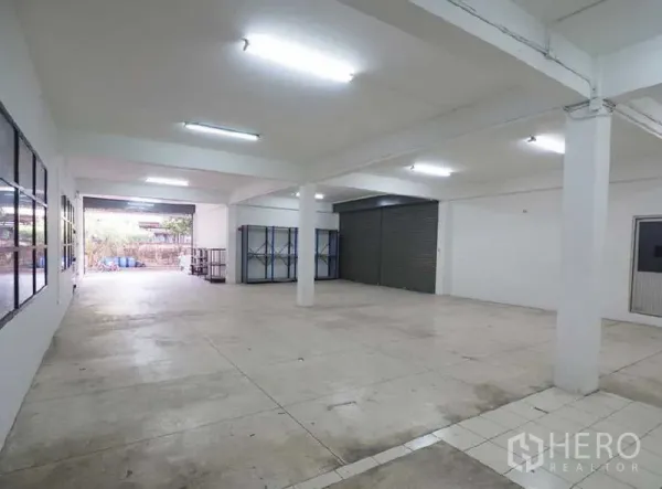 Warehouse for rent in Bang Kapi, Bangkok - Open warehouse bay with white columns, fluorescent lighting, concrete floor and a loading roller shutter.