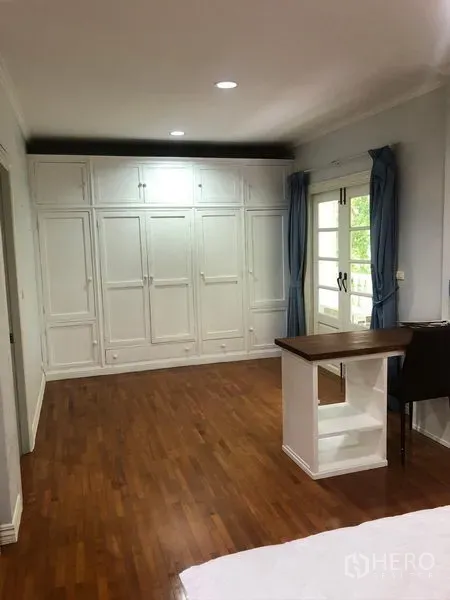 Townhouse for rent in Mueang Samut Prakan, Samut Prakan - Spacious bedroom with built-in white wardrobes, wood flooring, desk, and French doors with blue curtains.