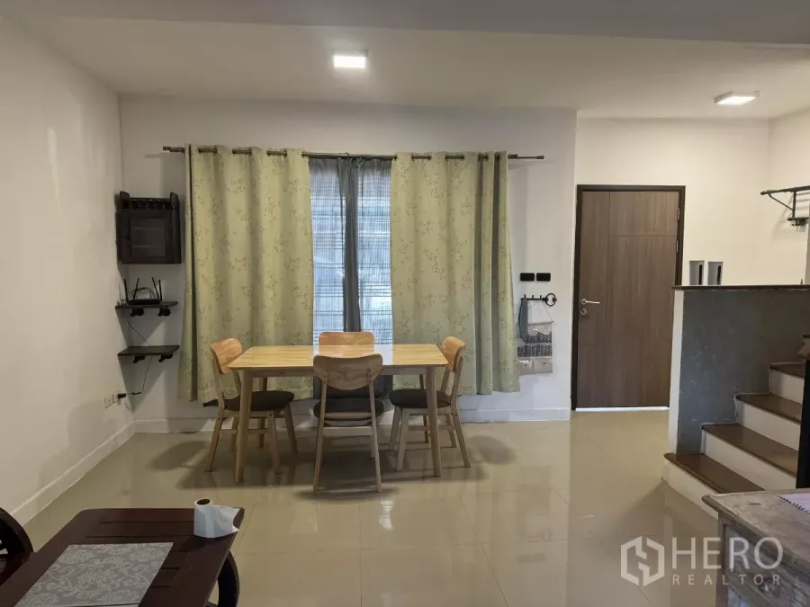 Townhouse for rent in Suan Luang, Bangkok - Bright dining nook with wooden table and four chairs, light curtains, side shelves, and tiled floor beside the entry door.