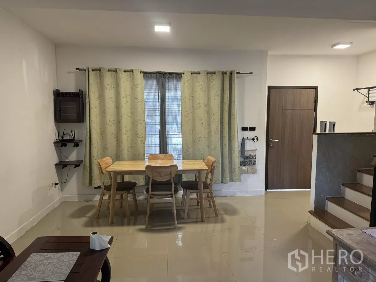 Townhouse for rent in Suan Luang, Bangkok - Bright dining nook with wooden table and four chairs, light curtains, side shelves, and tiled floor beside the entry door.