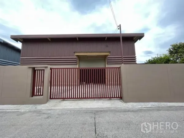 Warehouse for rent in Khlong Sam Wa, Bangkok - Front facade of the warehouse featuring a wide roller shutter door and secure red metal gate to the street.