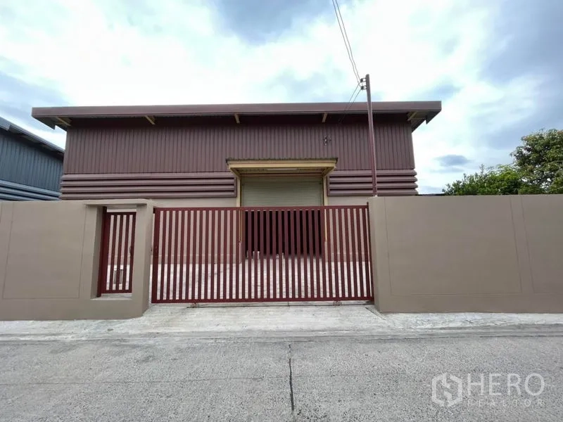 Warehouse for rent in Khlong Sam Wa, Bangkok - Front facade of the warehouse featuring a wide roller shutter door and secure red metal gate to the street.