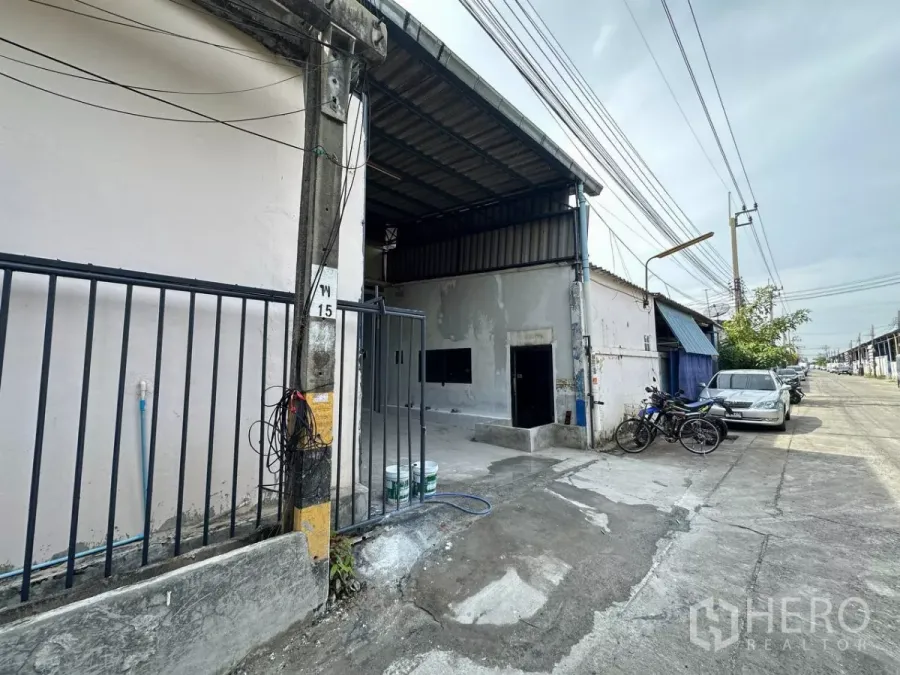 Factory for rent in Samut Prakan - Street-side view of the warehouse exterior with sliding gate, parking and overhead power lines in an industrial lane.