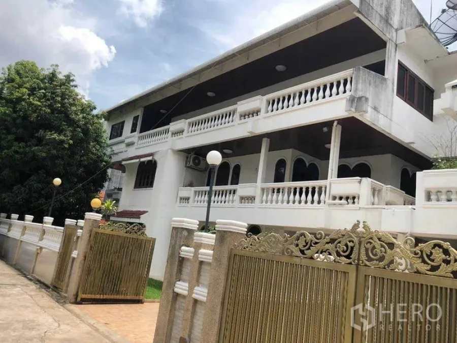 Detached House for rent in Watthana, Bangkok - Two‑storey detached house with white balustrades and ornate golden gate under a cloudy sky.
