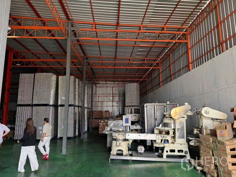 Factory for sale in Samut Prakan - High-bay warehouse with steel roof, stacked pallets and industrial machinery.