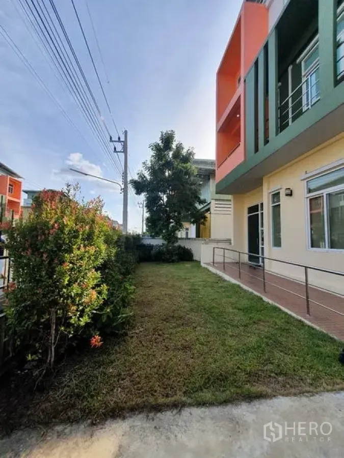Factory for sale in Lam Luk Ka, Pathum Thani - Modern factory exterior with green and orange facade, lawn, ramp and hedges along a quiet street.