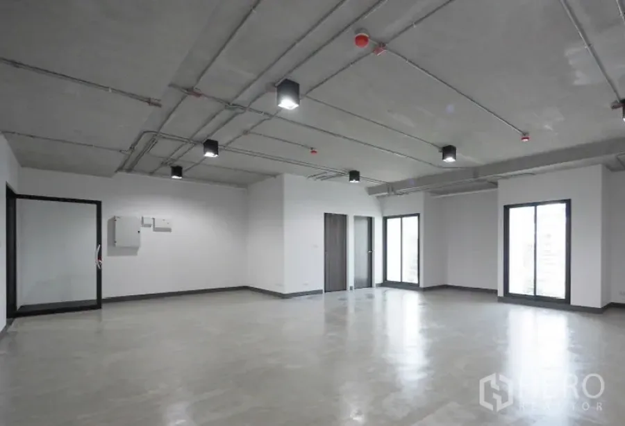 办公空间 出租在Khlong Toei, Bangkok - 明亮空置办公室，玻璃隔断入口与设备面板，工业风外露天花与多扇落地窗。