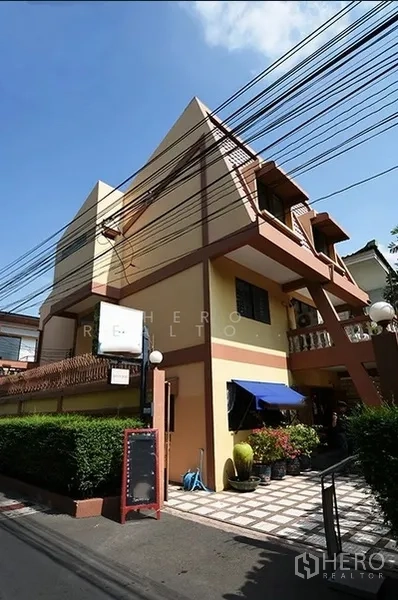 Detached House for rent in Watthana, Bangkok - Angular three-storey detached house in Watthana with balconies, an awning and a tiled entrance along a quiet lane.