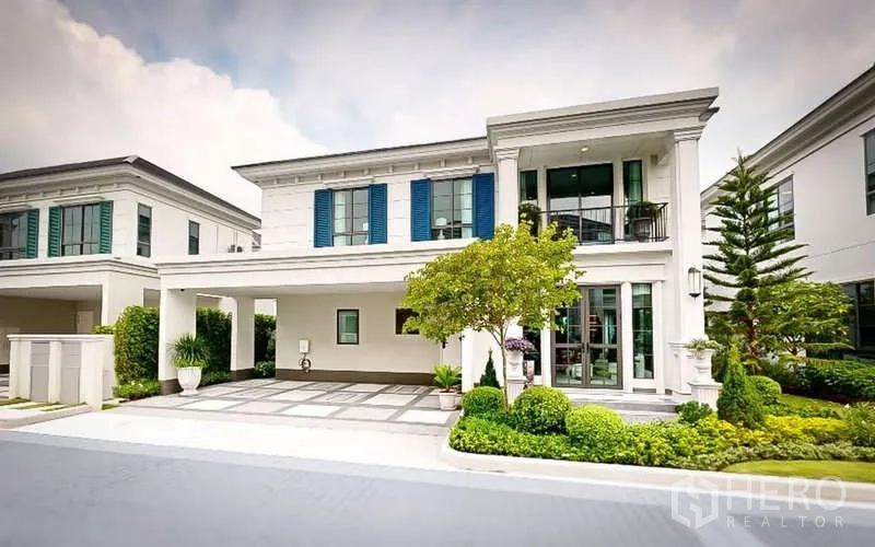 Detached House for rent in Bang Phli, Samut Prakan - Elegant two-storey detached house exterior with covered carport, blue shutters and manicured trees in a gated community.