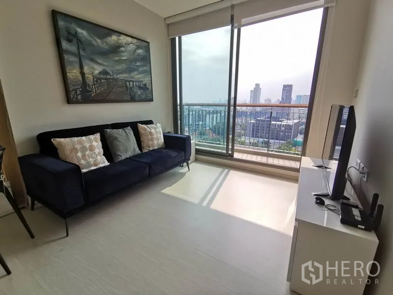 Condominium for rent in Khlong Toei, Bangkok - Living room with navy sofa and full-height glass opening to a balcony with Bangkok skyline.