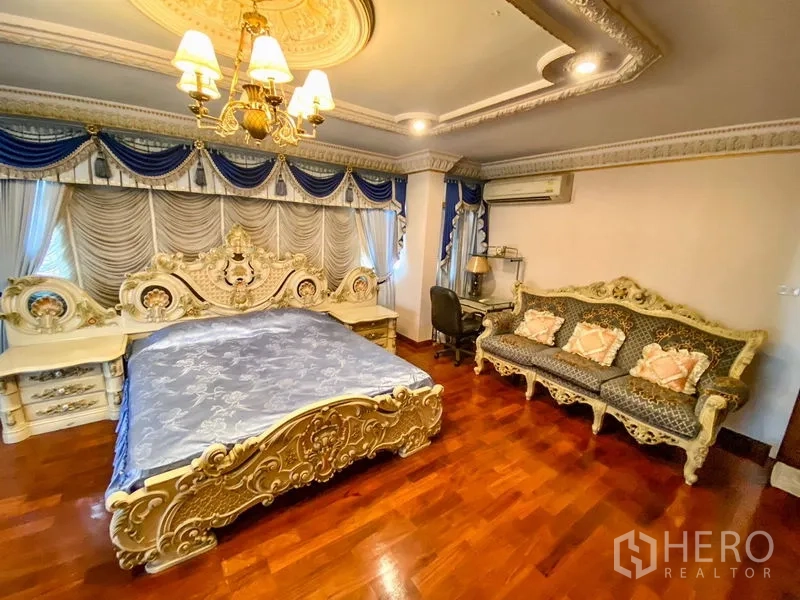 Detached House for rent or sale in Bang Khen, Bangkok - Large bedroom with blue drapes, carved bed, matching sofa, and wood floors.