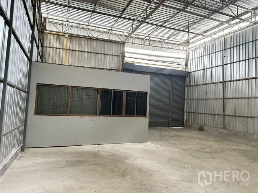 Warehouse for rent in Bang Khun Thian, Bangkok - Warehouse corner featuring an internal glass-partition office and large roller shutter door beneath a tall truss roof.