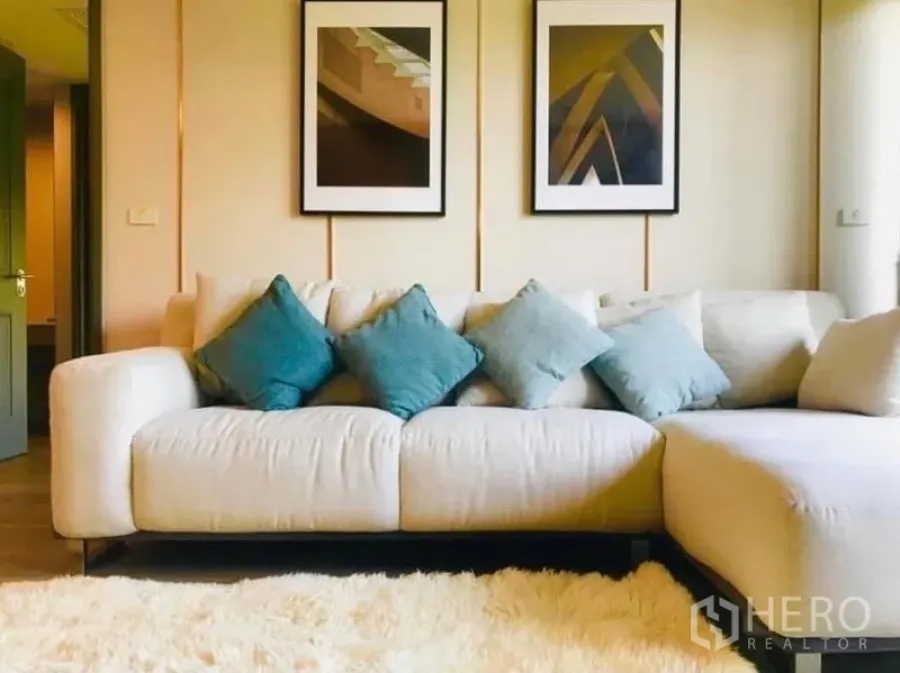 Condominium for rent in Pathum Wan, Bangkok - Cozy living room with cream L-shaped sofa, teal cushions, shaggy rug and framed art.