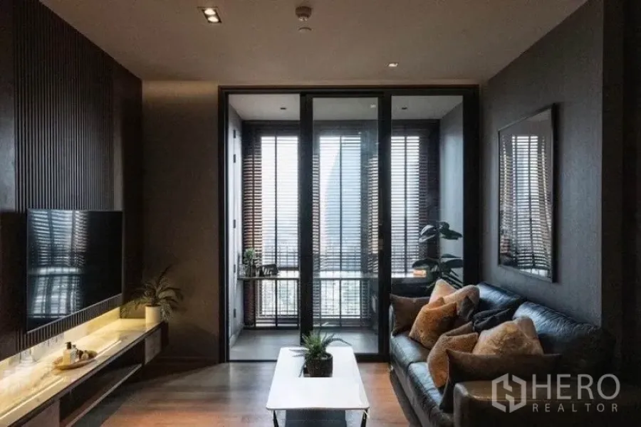 Condominium for rent in Khlong Toei, Bangkok - Modern living room with dark wood TV wall, leather sofa and glass doors opening to a bright sunroom with blinds.