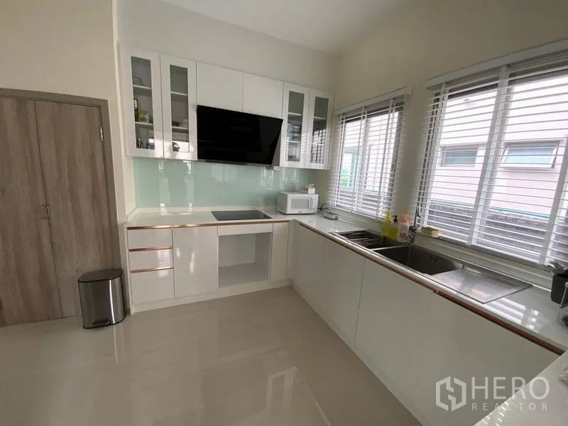 Townhouse for rent in Prawet, Bangkok - L-shaped modern kitchen with white cabinets, windows, sink and appliances.
