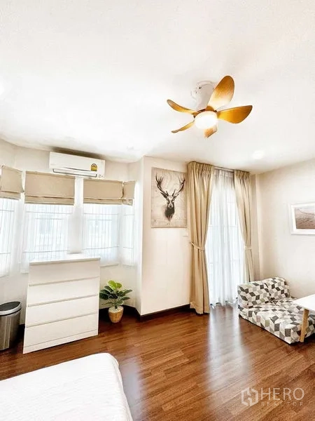 Townhouse for rent in Phra Khanong, Bangkok - Cozy bedroom with bay windows, gold ceiling fan and a small seating corner.