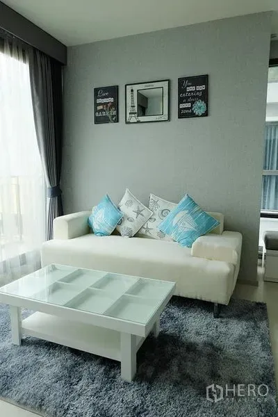Condominium for rent in Khlong Toei, Bangkok - Bright living room with white sofa, glass coffee table and full-height curtains.