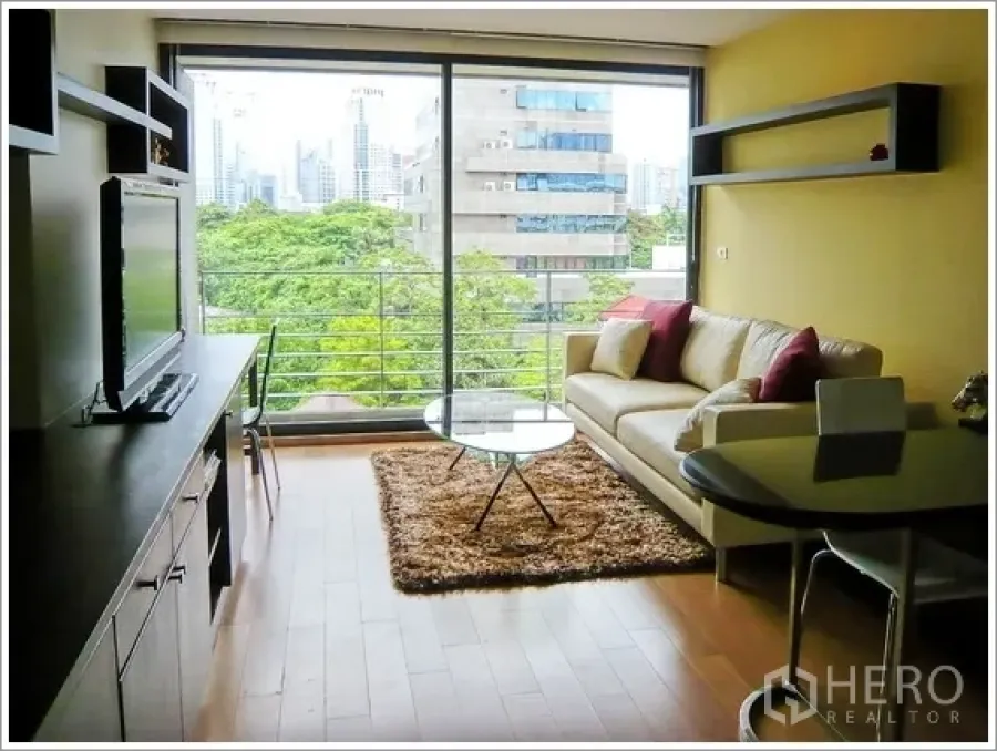 Condominium for rent or sale in Pathum Wan, Bangkok - Bright living room with floor-to-ceiling windows, balcony rail, wood floors, and a green park and skyline view in a Pathum Wan condo.