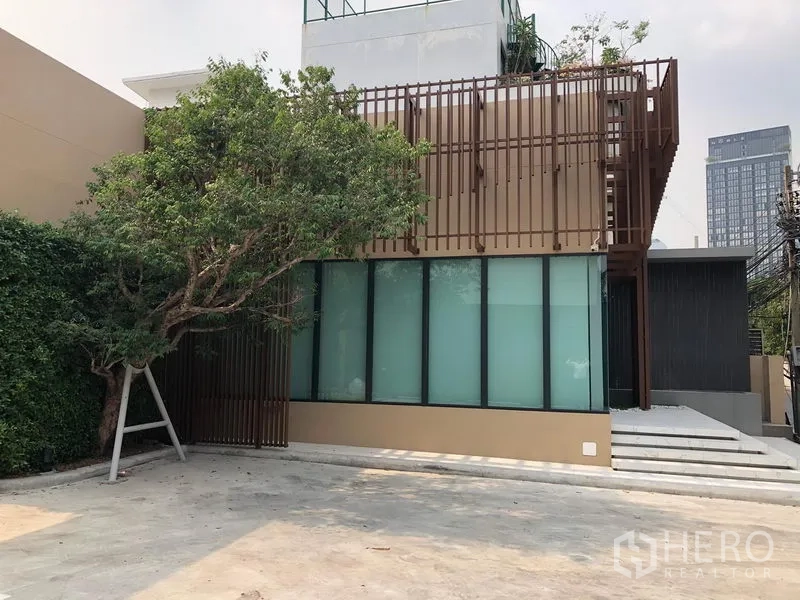 Retail Space for rent in Watthana, Bangkok - Street-level view of a contemporary retail unit with tree-shaded frontage, steps to the entrance and a wide concrete parking apron.