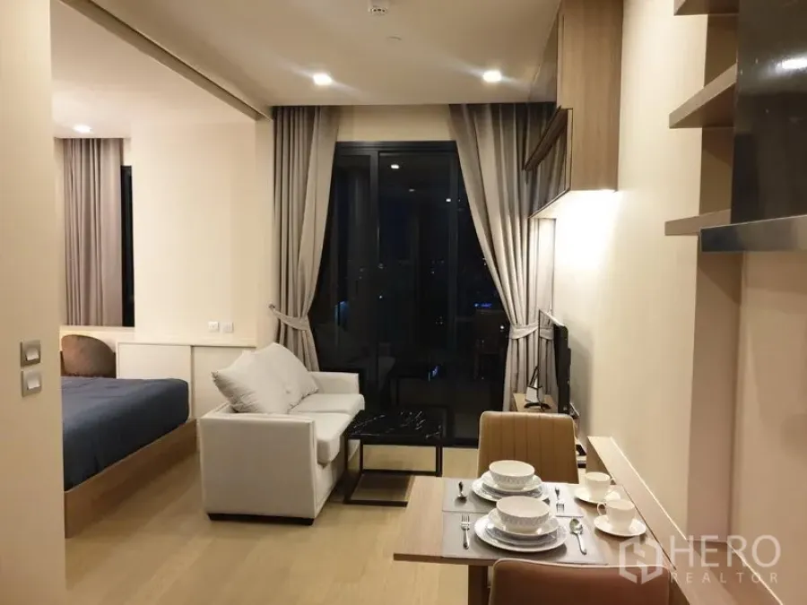 公寓 出租在Watthana, Bangkok - 开放式起居区，配沙发、电视与阳台落地门，左侧为睡眠区，夜景可见