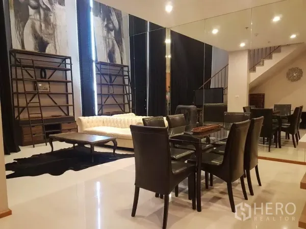Condominium for rent or sale in Khlong Toei, Bangkok - Dining area with glass table, mirrored wall and sofa in a double-height living room.