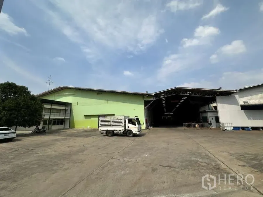Warehouse for rent in Pak Kret, Nonthaburi - Spacious concrete yard with green and white warehouse buildings, open loading bay and small truck.