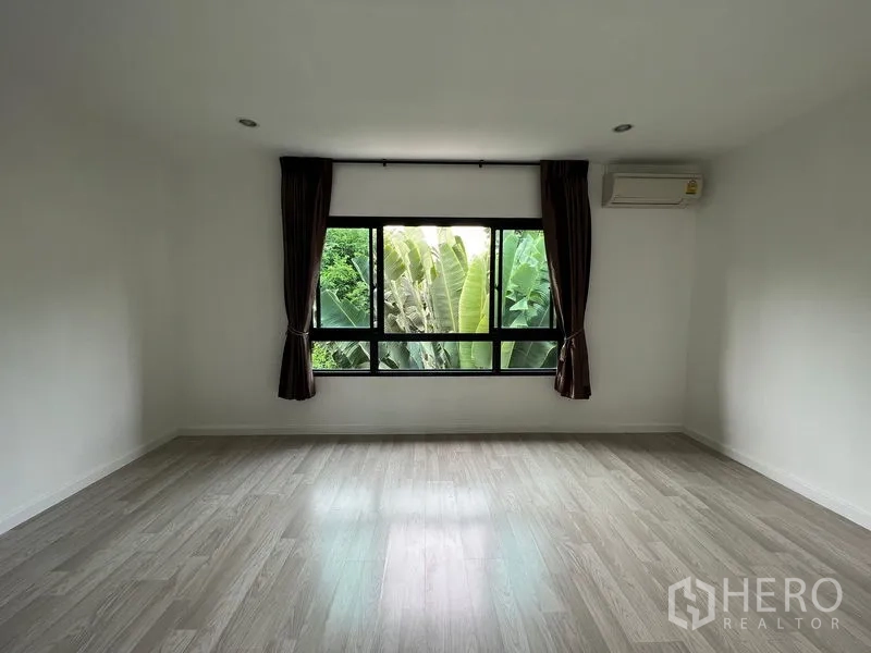 Townhouse for sale in Bang Kapi, Bangkok - Large bedroom with wood floors and wide window facing lush greenery.