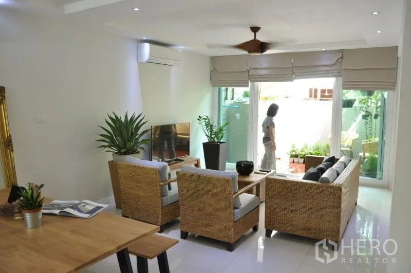 Townhouse for rent or sale in Watthana, Bangkok - Living area with woven furniture and TV opening to a terrace garden through sliding glass doors.