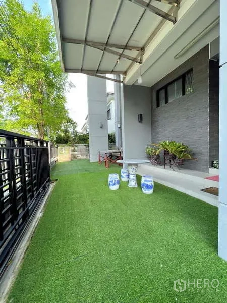 Detached House for sale in Prawet, Bangkok - Side yard with artificial grass lawn, covered patio, and potted plants along the fence.