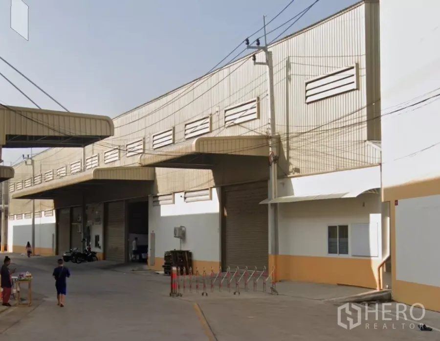 Warehouse for rent in Bang Khun Thian, Bangkok - Exterior view of a metal-clad warehouse row with multiple roll-up doors, covered loading bays, and a wide driveway in an industrial compound in Bang Khun Thian, Bangkok.