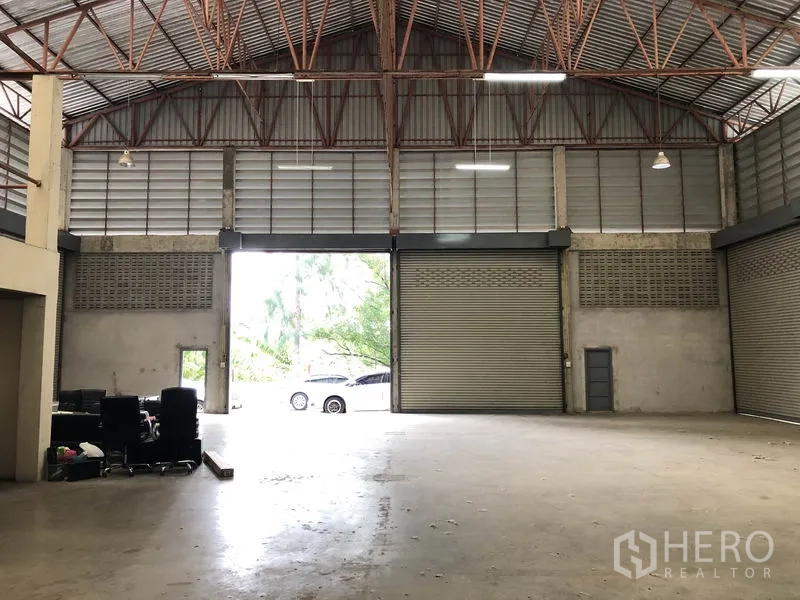 Warehouse for rent in Khlong Sam Wa, Bangkok - Interior view toward the entrance with two tall roller shutters and wide loading area; vehicles visible outside.