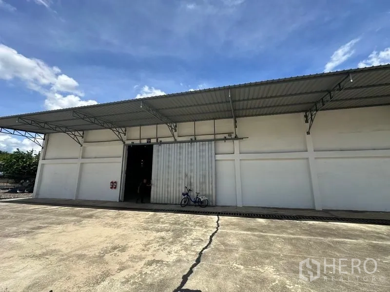 Warehouse for rent in Mueang Pathum Thani, Pathum Thani - Front exterior of a Pathum Thani warehouse with wide paved loading yard, large sliding metal doors and a canopy roof under blue sky.