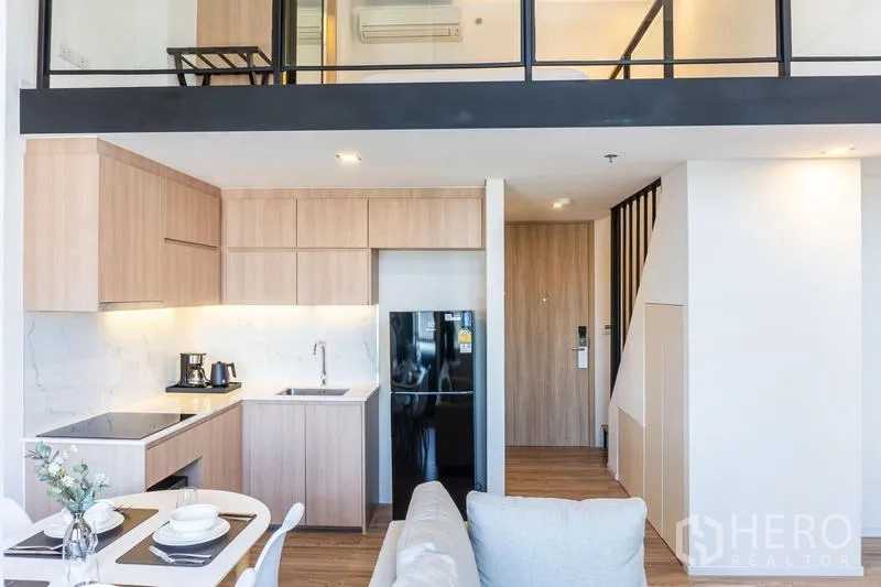 Condominium for rent in Khlong Toei, Bangkok - Open‑plan kitchen and dining under a loft mezzanine with wood cabinetry.