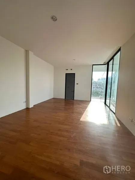 Detached House for rent in Watthana, Bangkok - Sunlit room with warm wooden floors and full-height sliding glass doors opening to a balcony with city views.