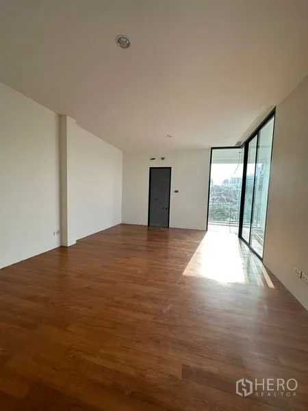 Detached House for rent in Watthana, Bangkok - Sunlit room with warm wooden floors and full-height sliding glass doors opening to a balcony with city views.