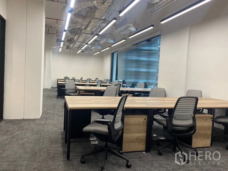 Office Space for rent in Pathum Wan, Bangkok - Open-plan office with long wooden desks, ergonomic chairs, carpet flooring, linear LED lighting and a large window.
