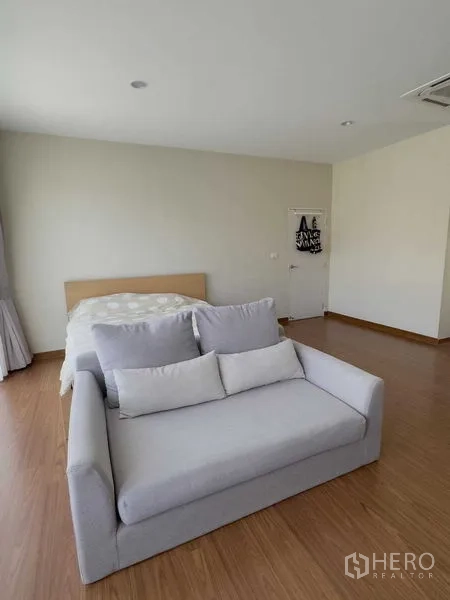 Detached House for rent or sale in Bang Khen, Bangkok - Master bedroom angle showing a gray loveseat at the foot of the bed and wood flooring.
