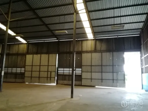 Warehouse for rent in Lam Luk Ka, Pathum Thani - Spacious warehouse interior featuring a high steel-truss roof, skylight strips, concrete floor, and oversized sliding doors.