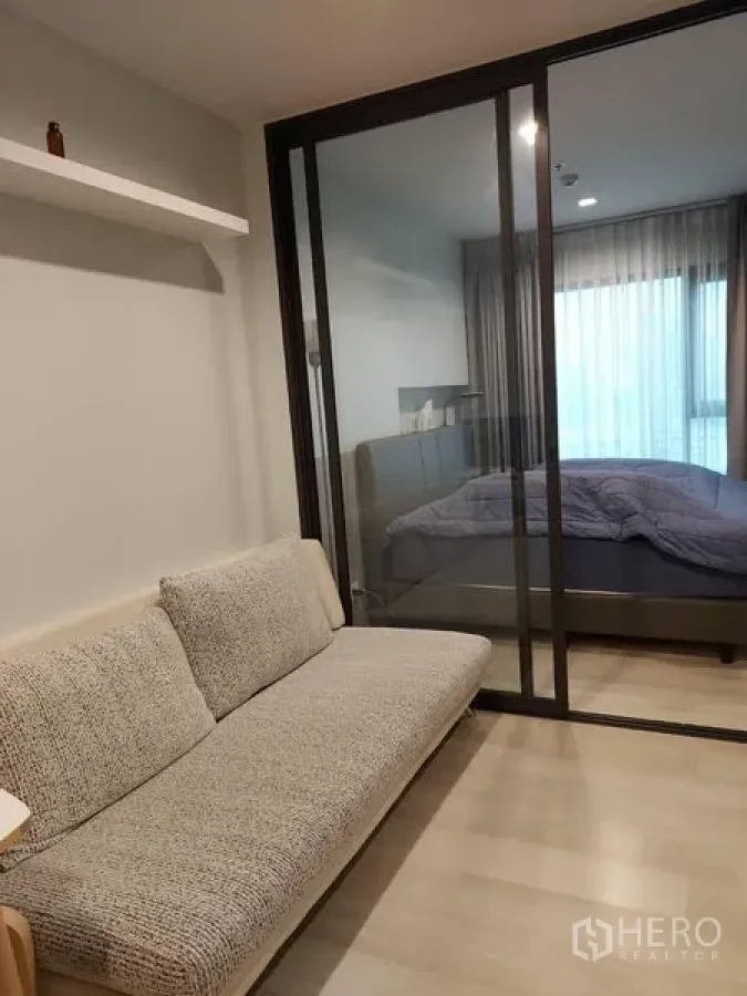 Condominium for sale - Living room with grey sofa and glass sliding door opening to a bright bedroom with floor‑to‑ceiling curtains.