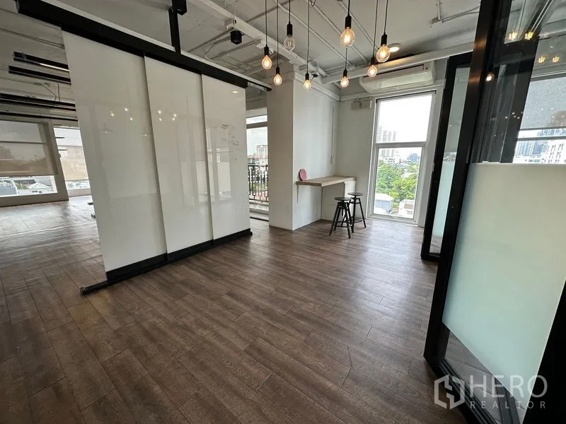 Office Space for rent in Watthana, Bangkok - Bright corner of an open‑plan office with wood-look floors, glass partitions, pendant lights, a built-in counter, and city views through tall windows.